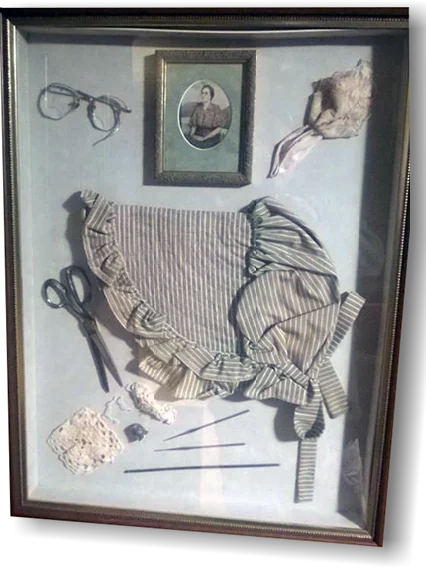 A shadow box framing an antique striped bonnet and apron with spectacles, sewing scissors, a crochet square and an oval portrait photograph.